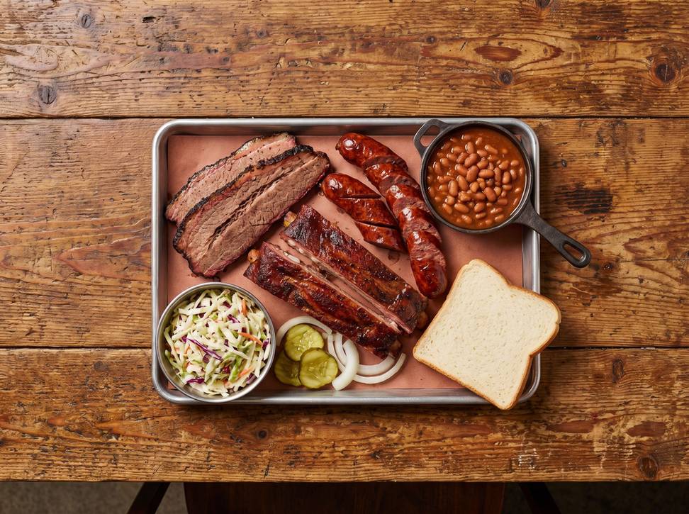 Overhead shot of a metal tray with sliced brisket, ribs, sausage, coleslaw, pickles, onions, beans in a cast iron pot and a slice of white bread